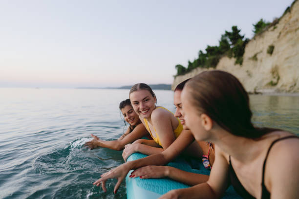 Four young girls are swimming in the ocean together. All of the girls are hanging on the surfboard as they look at one another and smile.