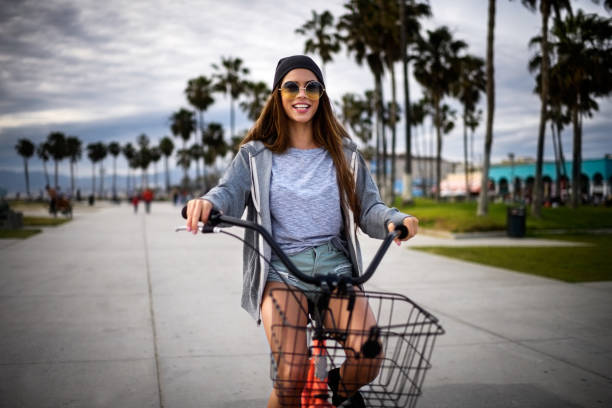A young woman is riding a bike alongside the boardwalk of a beach. She is wearing sunglasses and a beanie as she smiles and rides the bike.