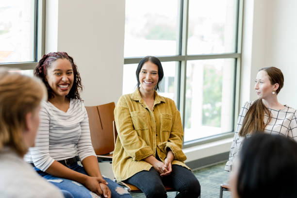 5 young women are sitting aside each other in a circle as they take part in a support group. All of the women are smiling and speaking to one another.