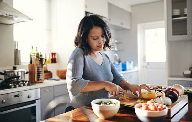 A young woman is standing in her kitchen cooking food. She is chopping vegetables on a chopping board.