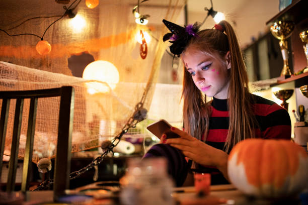 A young blonde teenage girl is sitting in a Halloween decorated room. She is dressed as a witch and has a tiny witch hat on her head. She is holding her phone in her hand as she scrolls on social media.