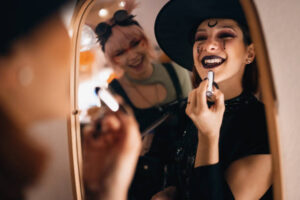 Two young girls are looking in the mirror as they prepare for a Halloween party. Both girls are dressed in Halloween costumes, as one of them applies black lipstick on her.