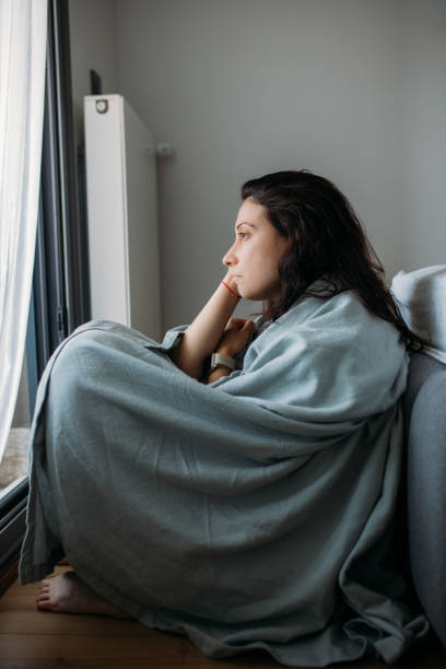 A young sad woman is sitting on the floor of her bedroom. She is looking out of her glass door, as she is covered by a blanket.