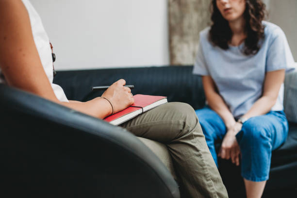 In this image we see a client in her therapy session. She is sitting across from her therapist on a couch. Both women are facing one another, as the therapist holds her notebook on her lap.