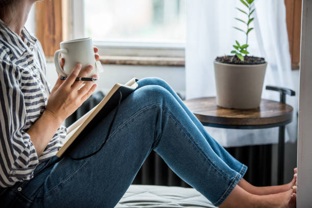 A woman is sitting on a couch with her feet leaning on the wall. She is holding a cup of coffee is her hands, as her journal is on her lap. She is also holding a pen in her hand.