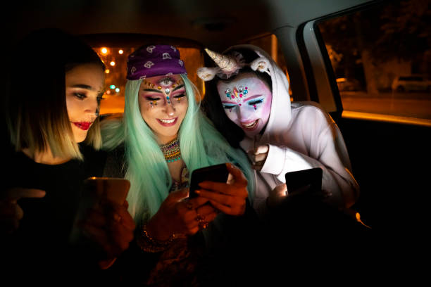 Three young teenage girls are sitting in the back of a car beside each other. They are all dressed up in costumes for a Halloween party and they all have face paint on. They are all holding their phones in their hand.