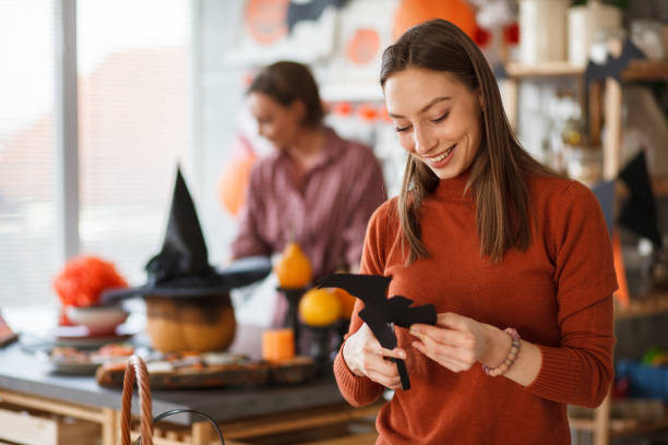 A young woman is preparing for a Halloween party as she cuts out a bat from black construction paper. Behind her is a pumpkin with a witch hat on it.