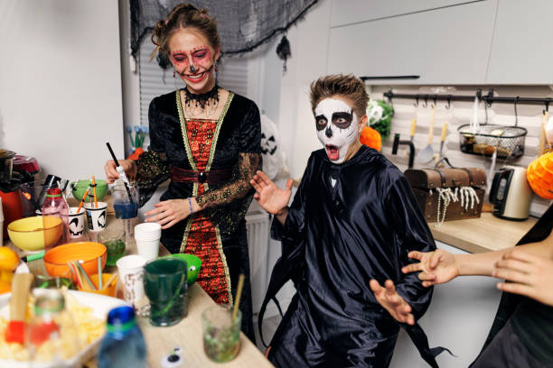 Two young teenagers are at a Halloween party standing in the kitchen. The teenage girl is dressed up as a witch beside a teenage boy who is dressed as a skeleton.