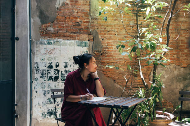 A young Asian American woman is sitting in an outdoor cafe surrounded by beautiful greenery. She is leaning her chin on her hand, with a pen in her other hand. In front of her on the table is an open journal.