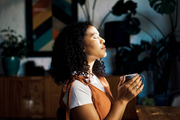 A biracial woman is standing in her home enjoying the sunlight that is shining on her face. She has her eyes closed and is holding her coffee cup in her hands.