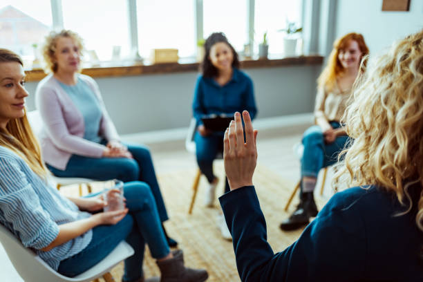 In this image we see group therapy happening with 4 woman members. The group leader is also a woman who is holding her hand up as she faces the rest of the group.