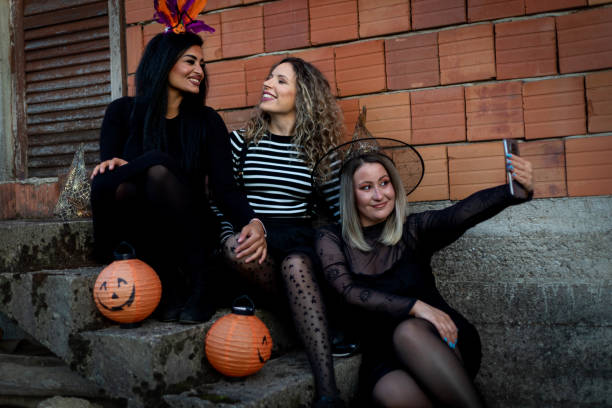 Three young women are sitting on the steps outside of a house. They are all dressed up in Halloween costumes and are taking a selfie together.