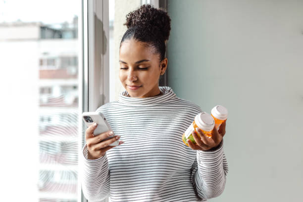 An African American woman is standing in her home, leaning on the window pane as she holds 2 medication bottles in one hand. In her other hand she has her phone.