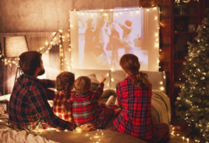In this image we see a husband and wife with their two children sitting in between them on the bed. The house is decorated in Christmas decorations, as they watch a Christmas movie on a projector.