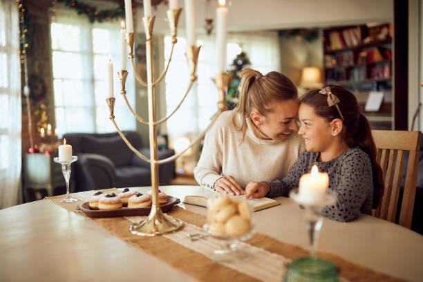 A mother and daughter are sitting at their dinner table in their home. On the table there are cookies and a menorah. They are reading a book together.