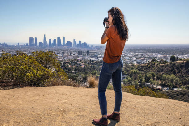 A young woman is standing on a hike path and taking photos with a professional camera of the Los Angeles skyline.