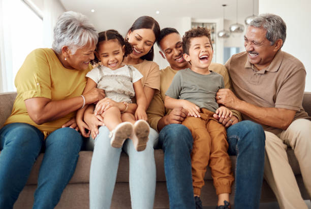 In this image we see a family of 6 sitting beside each other on the couch. The two grandparents are sitting on the outer ends of the couch, as the two young parents sit in the middle with their two children on each of their laps. They are all laughing together. Parenting Tips