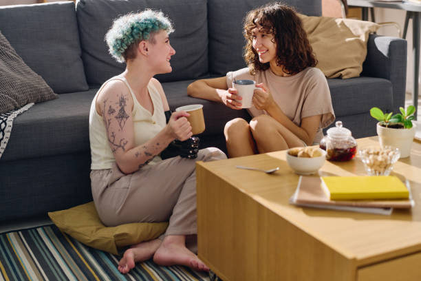 A young LGBTQ couple is sitting together in their living room on the floor. One of the partners has a prosthetic arm and is holding a coffee mug.