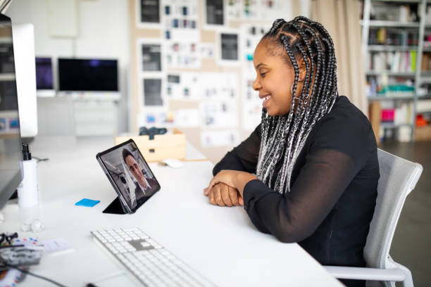 A young African American woman is sitting in her office at her desk. She is doing a video meeting on her tablet with a young American man.