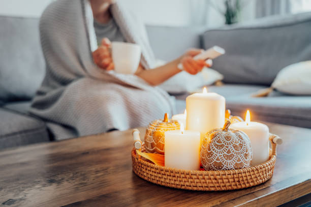 In this image we see a zoomed in view of table decor. There are lit candles and decorate pumpkins in a basket in the middle of a coffee table. A woman is sitting on the couch covered by a blanket and holding the remote and a coffee mug.