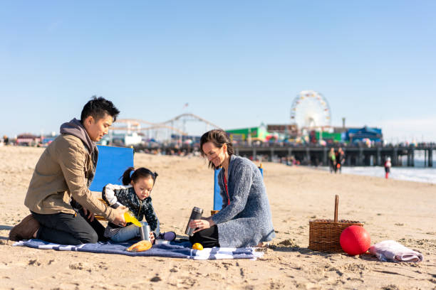An Asian American family of three are on a blanket at the beach enjoying a picnic. Behind them we see the Santa Monica pier. Parenting Tips