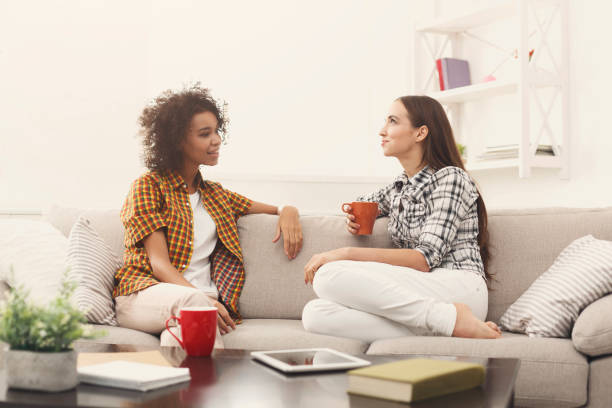 Two young women are sitting together on a couch. They are talking together and one of the women is holding a coffee mug in her hand.