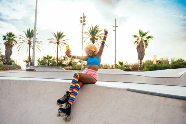 A young African American woman is sitting on a skating ramp. She is wearing rollerblades and is taking a selfie with her phone.