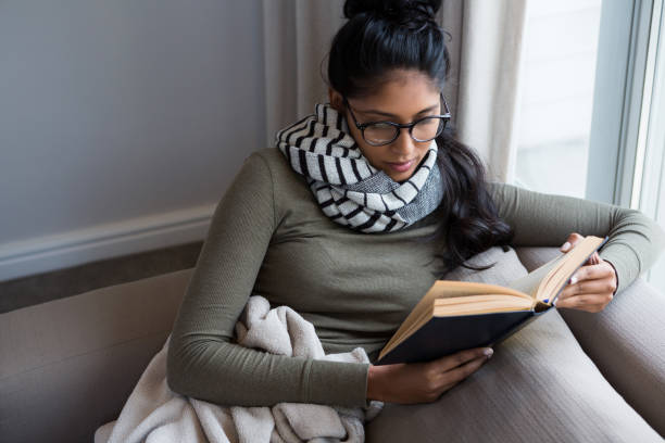 A young woman is sitting in her home on the couch reading a book. She is wearing a scarf and glasses, and is covered with a blanket.

balance during the holiday season