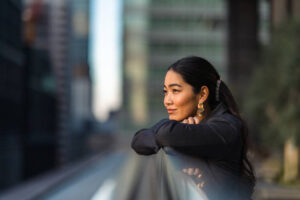 A young Asian American woman is standing outside in the city leaning on a ledge with her head on her hands.