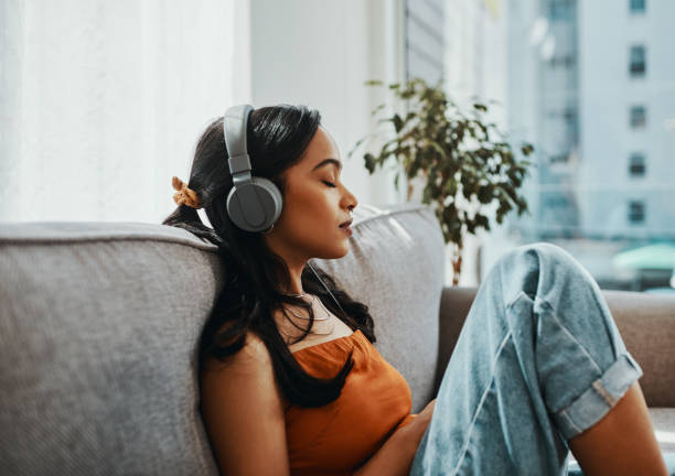 A young biracial woman is sitting on her couch with her knees propped up. She is wearing a headset while listening to music with her eyes closed.