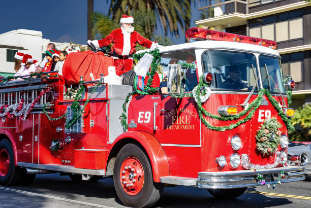 In this image we see a firetruck decorated with Christmas decorations. There is a Santa standing on top of the truck with a bag of gifts.