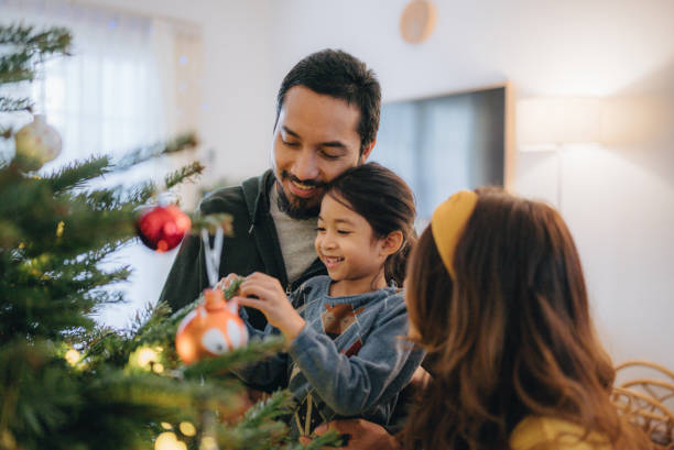An Asian American family of three is decorating their tree together. The father is holding their daughter as she puts an ornament on the tree.