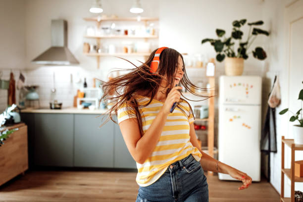 A young woman is dancing in her kitchen. She is wearing headphones and smiling as she dances.