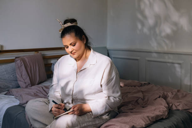 A young beautiful plus sized woman is sitting on her bed in her bedroom. She is writing in her journal with a pen. 