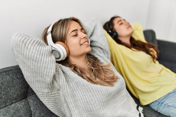 Two young women are sitting on a couch next to each other. They are both wearing headphones and listening to music with their eyes closed. They both have their arms behind their heads.