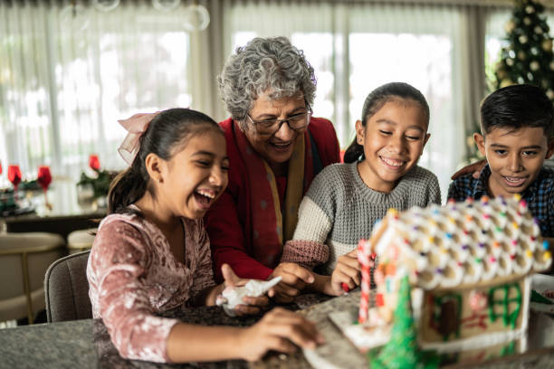 Three Hispanic children are decorating a gingerbread house with their grandmother. They are all smiling and happy.