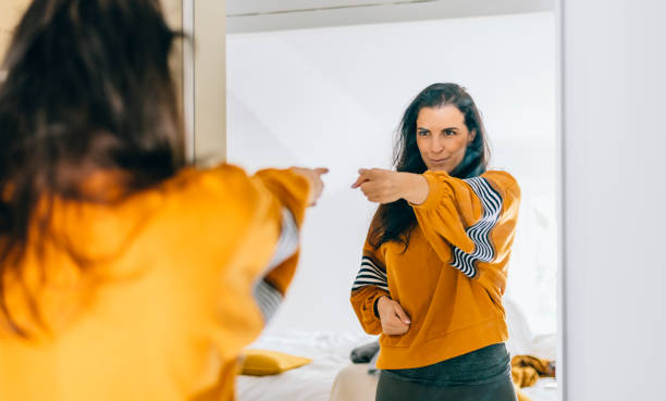 A young woman is looking at herself in the mirror of her bedroom. She is pointing at her reflection. She is wearing a bright yellow sweater.