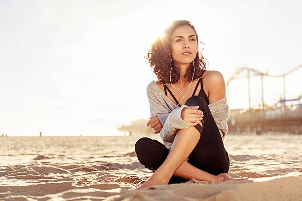 A woman is sitting on the sand at the beach. She is wearing headphones and listening to music. The Santa Monica pier is behind her in the distance.