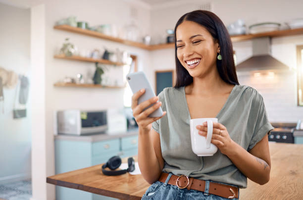 A young woman is standing in the kitchen of her home. She is holding a coffee mug in one hand and using her phone as she smiles and laughs.