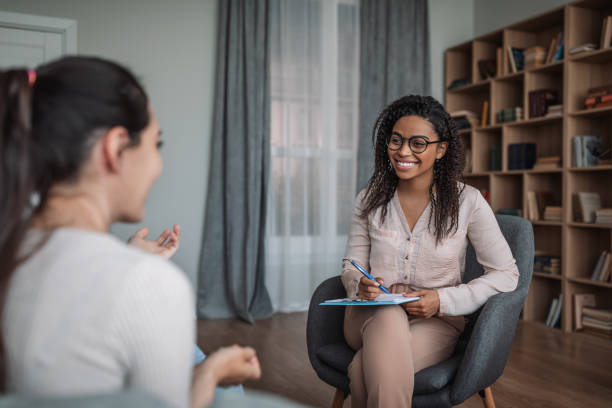 There are two young women in a therapist office. The therapist is a young African American woman sitting across from the other woman as she holds her clipboard.