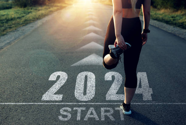 A woman is standing at the start line of a race. She is stretching her leg. On the ground it is written "2024 START"