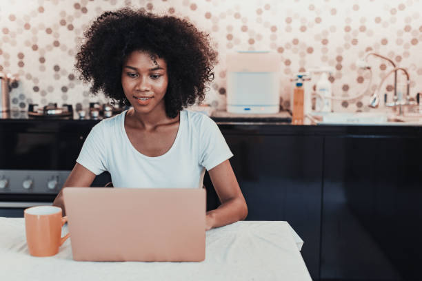 A young African American woman is sitting at her dinner table in her kitchen with her laptop open in front of her. She has a cup of coffee beside her laptop as she does work.