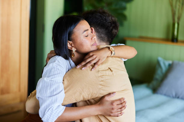 A biracial couple is sitting in their home, embracing each other. They are hugging tightly, and the woman has her eyes closed.