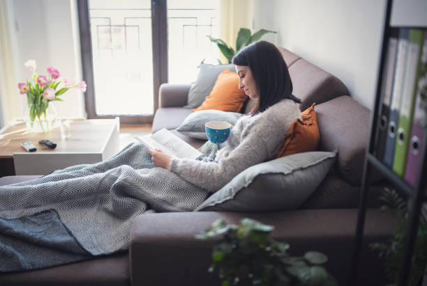A young woman is laying on her couch in her living room. She is covered by a blanket and is drinking coffee as she reads her book.