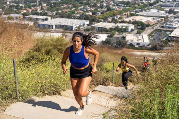 2 young women are running up a hiking trail of stairs. They are surrounded by greenery and are wearing workout clothing.