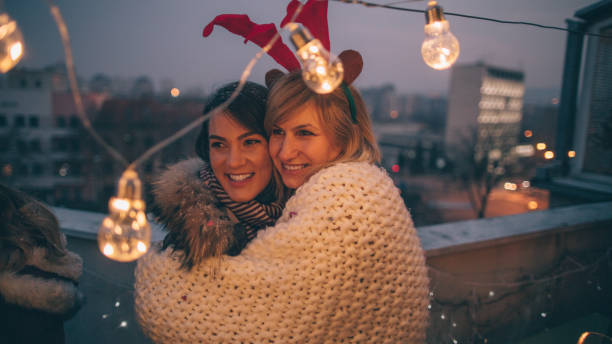 Two young women are hugging each other standing on a balcony. They are both smiling beside each other.