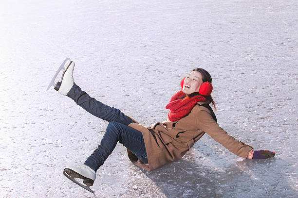 A young Asian American woman is fallen in an ice rink. She is wearing ice skates and ear muffs, as she smiles and raises her leg in the air.