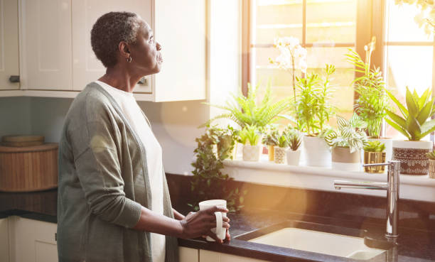 An older African American woman is standing in her kitchen by the sink. She is holding a coffee mug in her hands as she looks out the window.