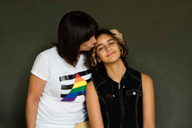 A woman is standing next to her non-binary daughter giving her a kiss on her head. The young individual is smiling as she leans into her mother.