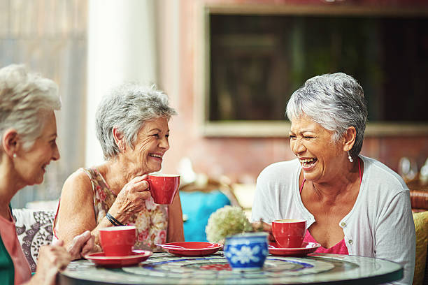 Three senior citizen women are sitting together at a cafe drinking coffee and laughing.